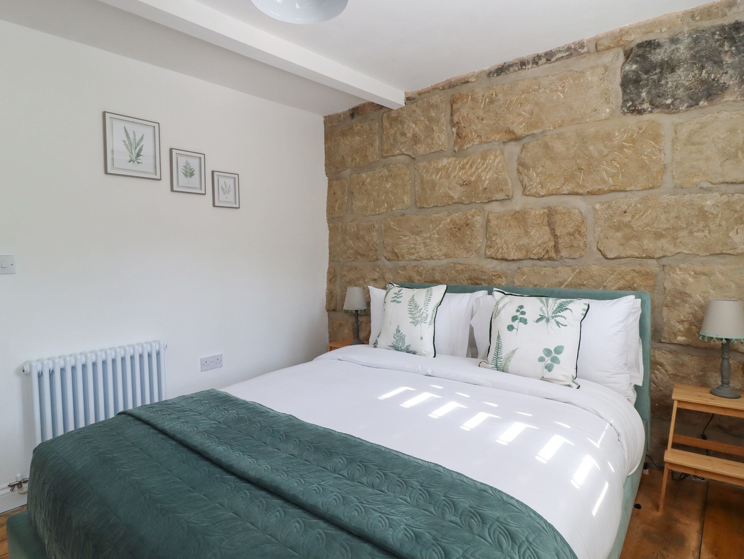 Quarto com parede de pedra, cama com roupa de cama verde e duas gravuras botânicas emolduradas na parede.