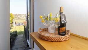 A bottle of champagne on a wooden shelf with a wicker basket, two clear glasses with flowers, and a tag that says "Enjoy". - Penistone View (Keighley)