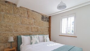 A bedroom with a stone wall, a bed with a green headboard, white bedding, and a window with blinds. - Penistone View (Keighley)