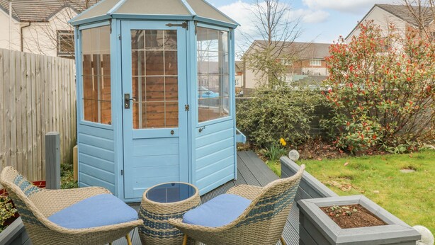 A blue wooden shed with a glass door, surrounded by a garden with a wooden fence, a wicker chair with a cushion, and a small table. - 10 Goodhope Gardens (Aberdeen)