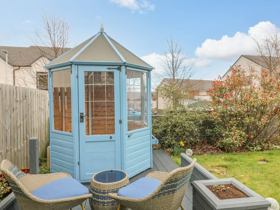 A blue wooden shed with a glass door, surrounded by a garden with a wooden fence, a wicker chair with a cushion, and a small table.