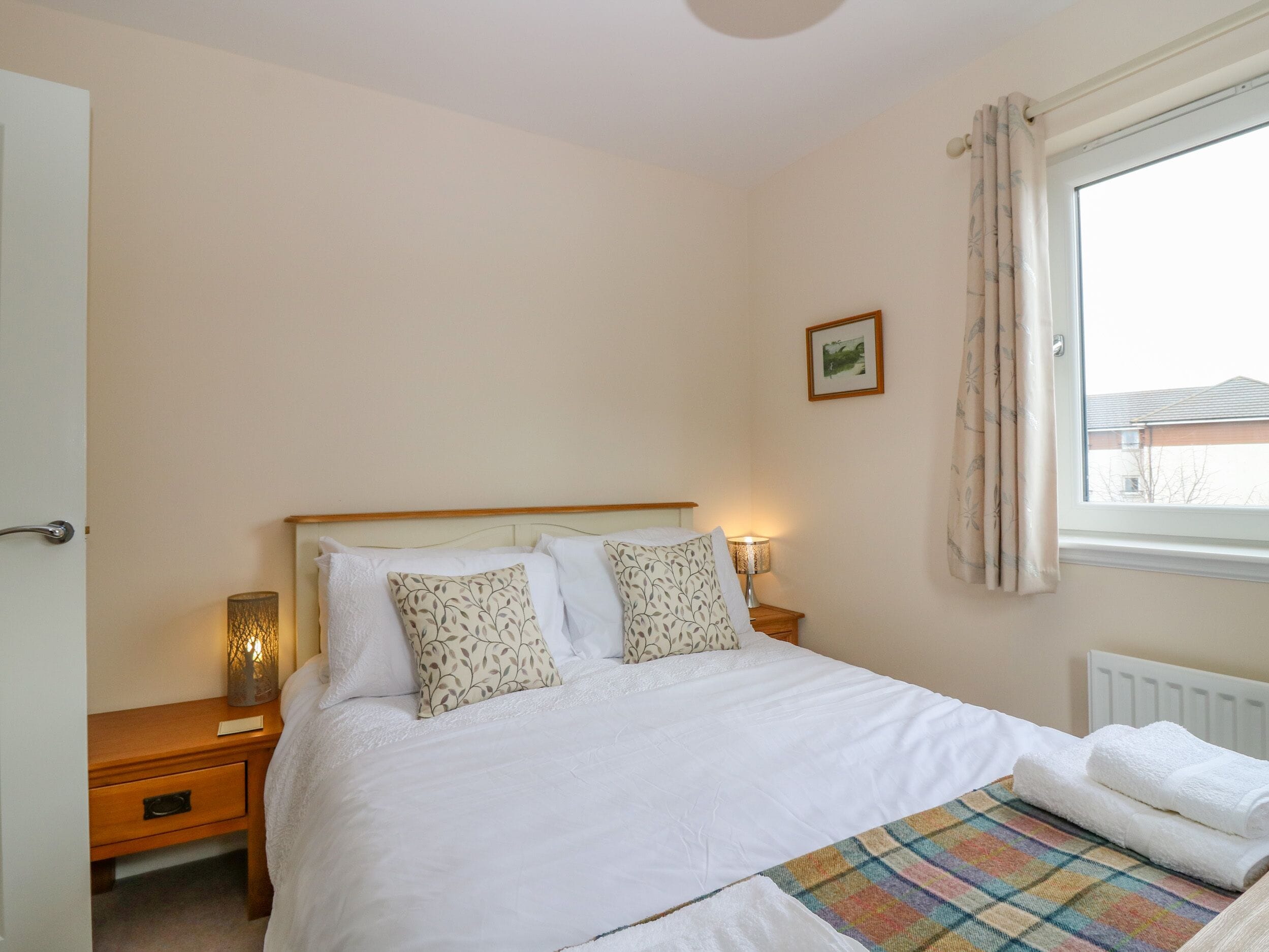 A neatly made bed with white bedding and patterned pillows, a bedside lamp, and a window with curtains.