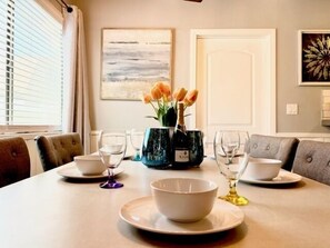 Una mesa de comedor servida con platos, tazones y copas de vino blancos, acompañada de un florero con tulipanes naranjas y una botella de champán.