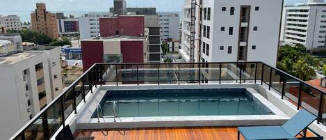 Una piscina en la azotea con cubierta de madera y sillones de descanso, rodeada de edificios de la ciudad y un cielo despejado.