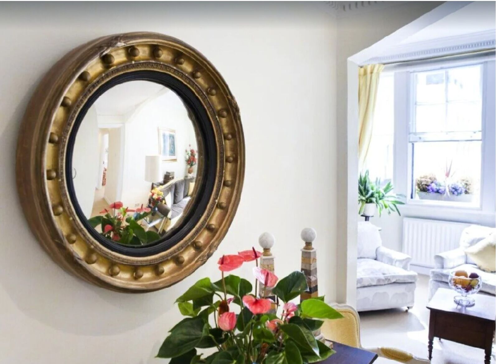 Een kamer met een ronde spiegel aan de muur, een witte bank, een tafel met bloemen en een raam met planten.