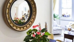 Una stanza con uno specchio tondo, un divano bianco, un tavolo con un vaso di fiori e una finestra con piante.