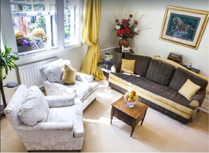 A living room with a sofa, armchair, and a small table with fruit. There is a window with curtains, a radiator, and a painting on the wall.