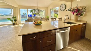 Una cocina moderna con una isla grande, lavavajillas de acero inoxidable y una zona de comedor con vista al océano.