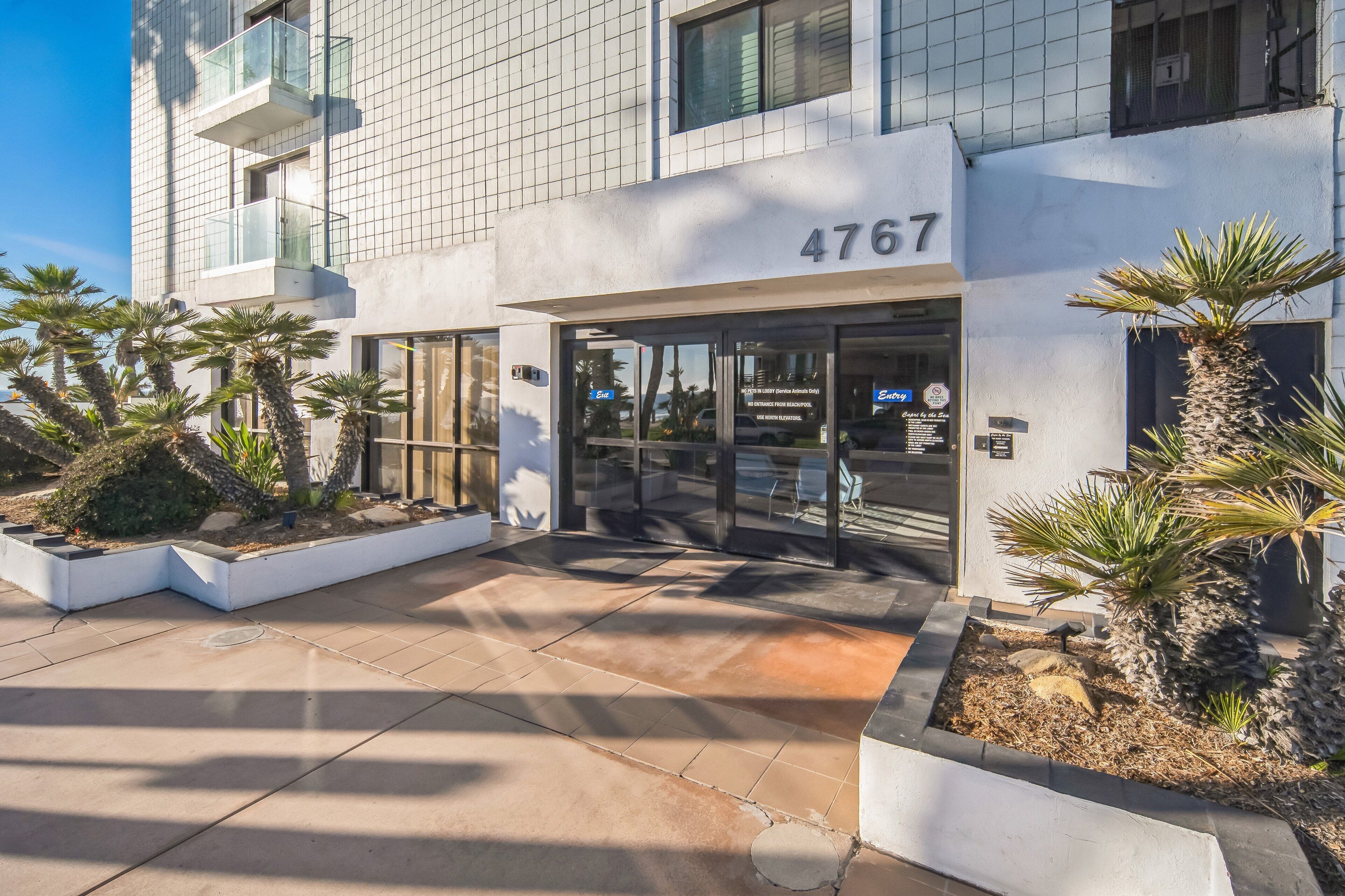 A modern building with the number 4767, featuring a glass entrance, outdoor seating, and palm trees.