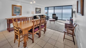 Un'ampia sala da pranzo con tavolo e sedie in legno, un divano e vista sull'oceano attraverso ampie finestre.