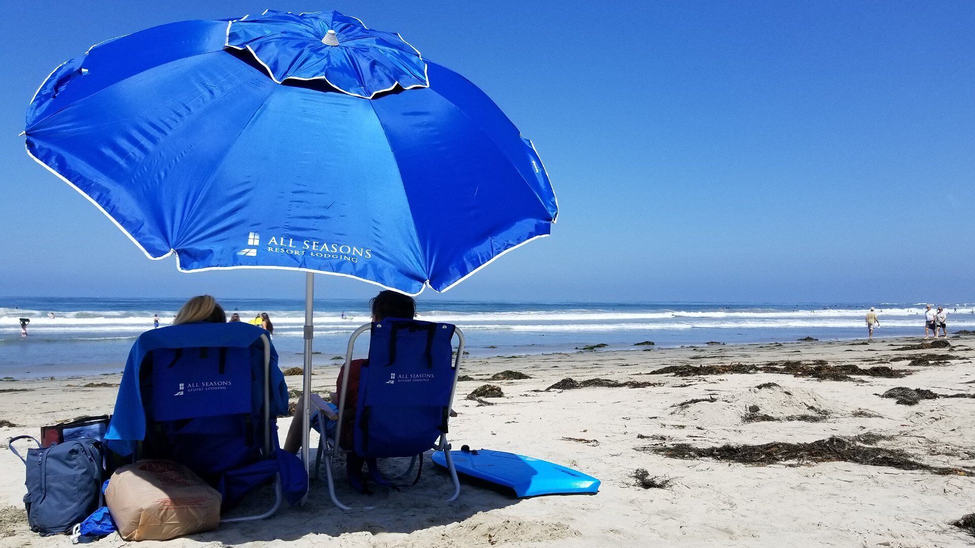Two beach chairs under a blue umbrella with 'All Seasons Resort Lodging' branding, on a sandy beach with ocean waves in the background.