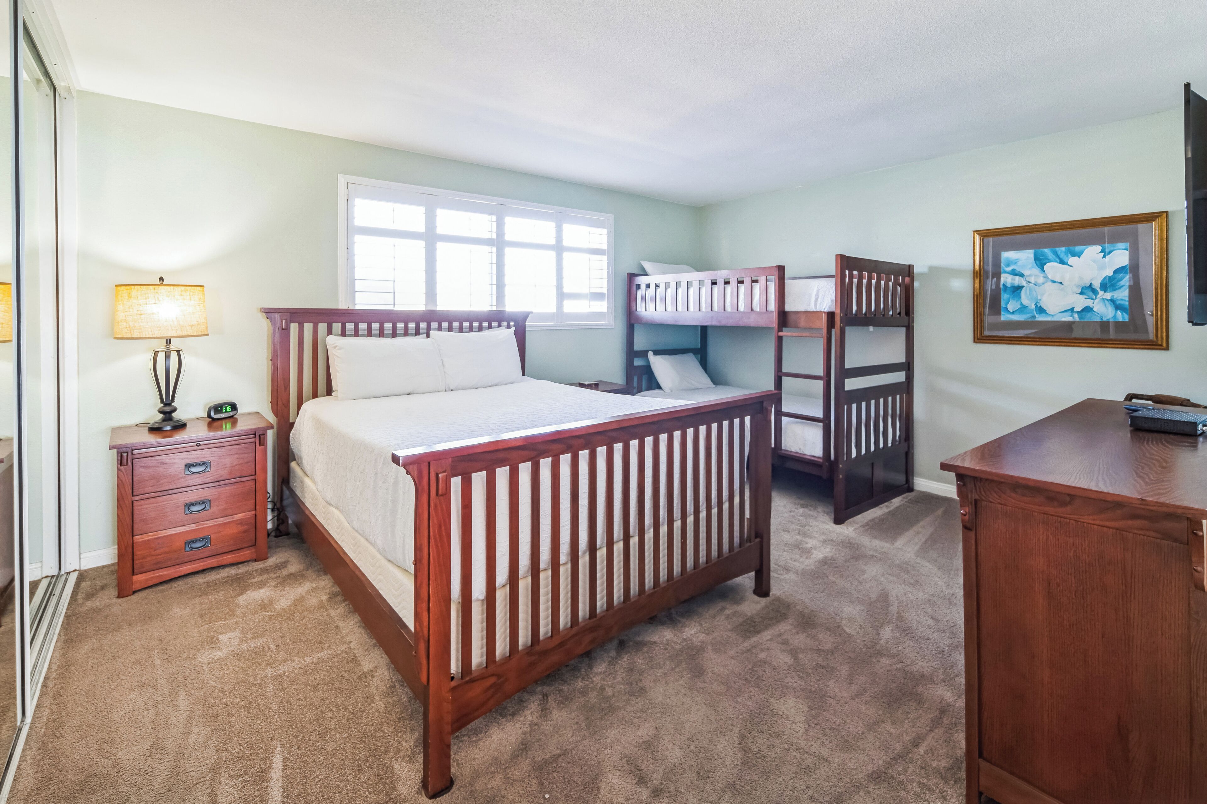 Een slaapkamer met een houten bed, nachtkastje, dressoir, stapelbed en een raam.