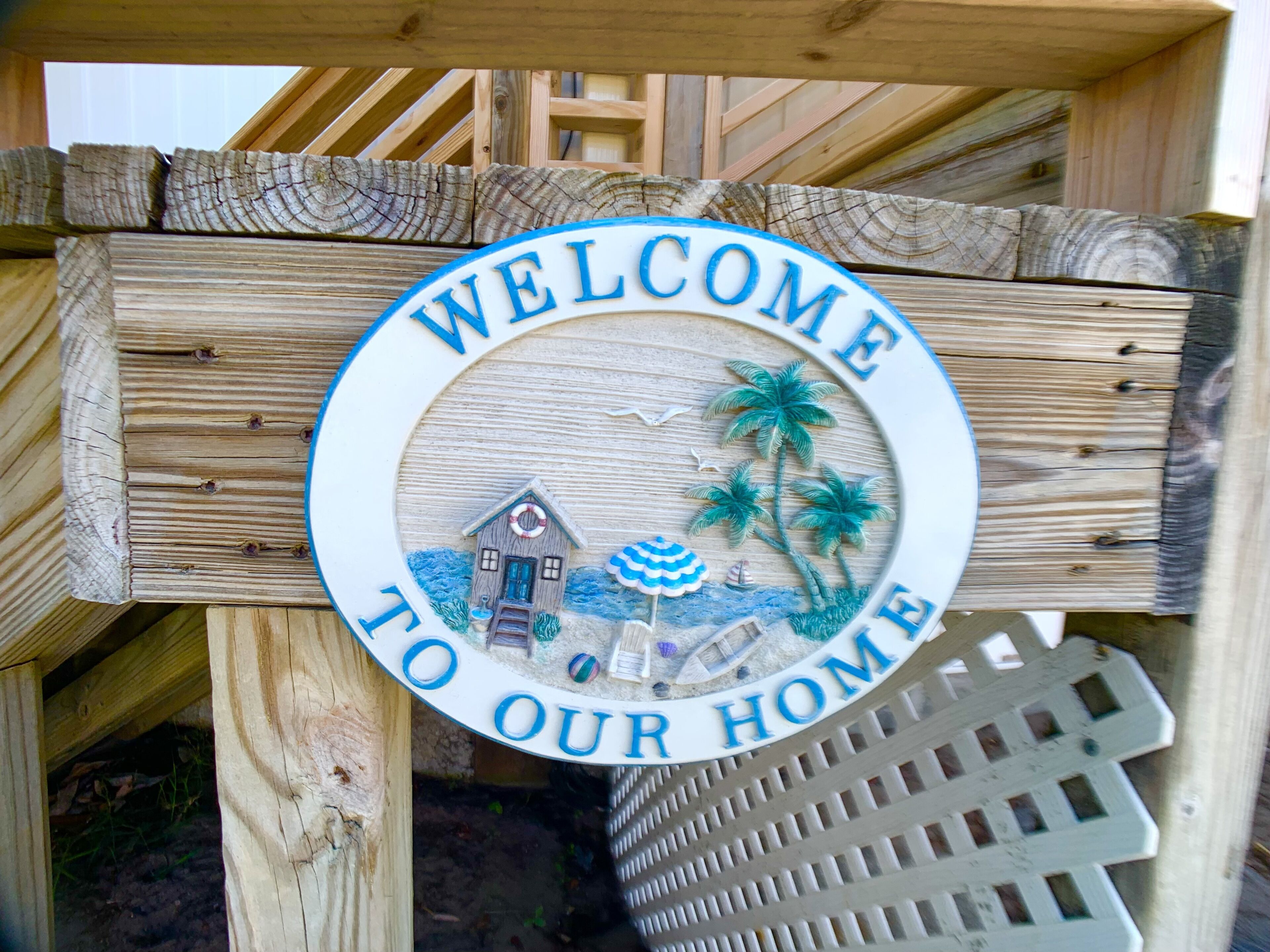 A wooden sign with a circular design, featuring a house, palm trees, and a beach scene, with the text "WELCOME TO OUR HOME".