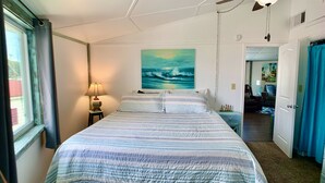 A bedroom with a bed, a nightstand, a lamp, a painting of waves, and a door leading to another room. - ❤️Amazing Round House - Easy Self Check-In, Just Steps to Beach! (Carolina Beach)