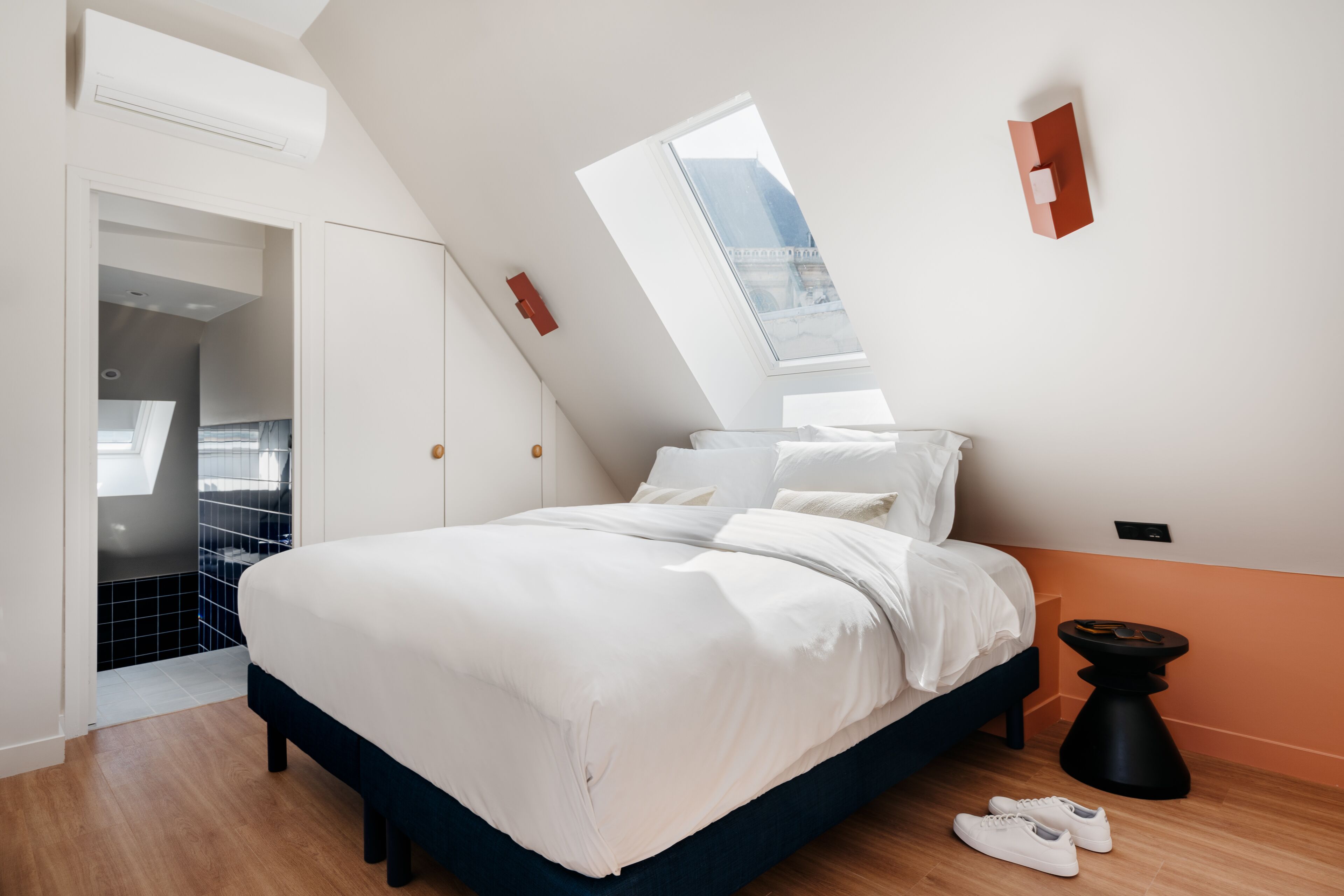 A bedroom with a sloped ceiling, a bed with white bedding, a black side table, and a window.