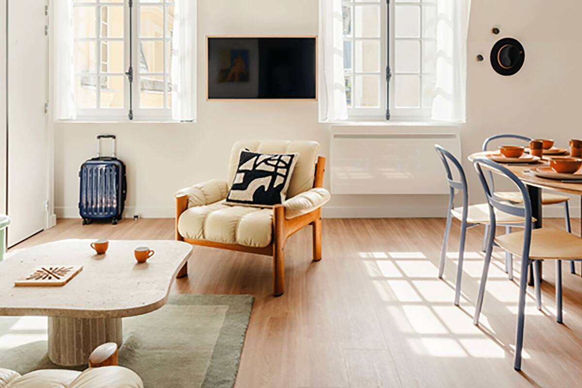 A cozy living room with a wooden sofa, a small round table, a dining table with chairs, a television mounted on the wall, and a window with curtains.