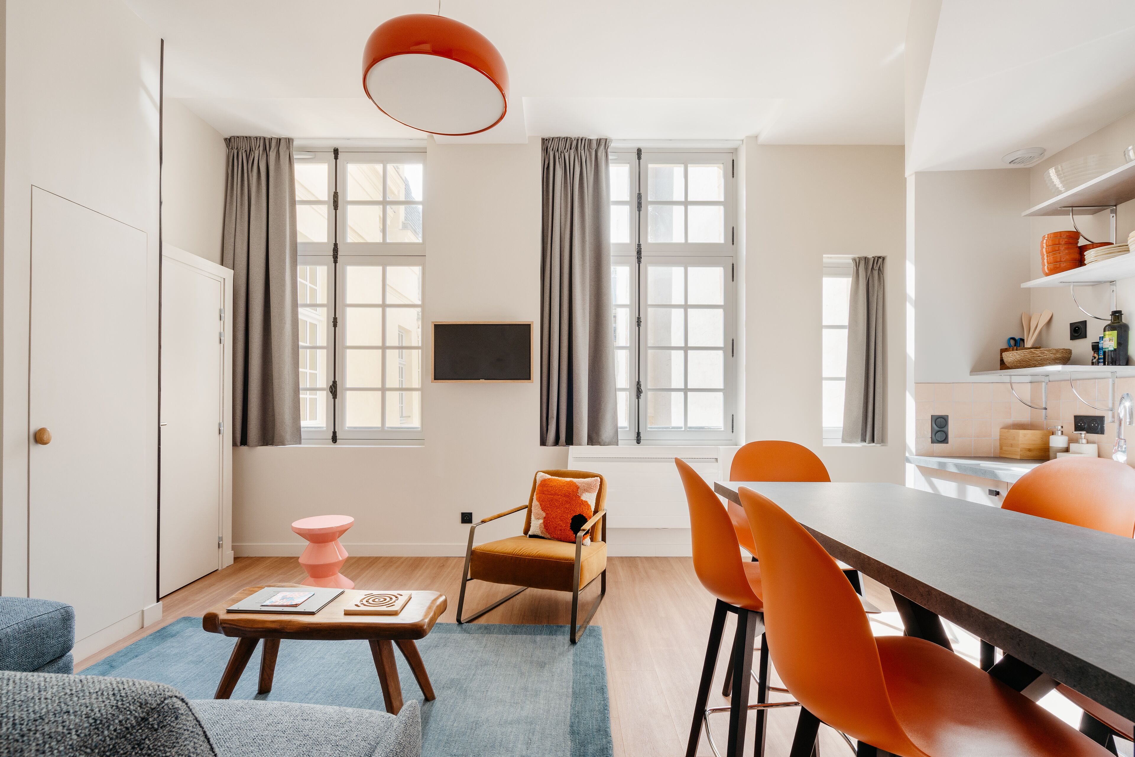 A modern kitchen with a dining area, large windows, and a flat-screen TV mounted on the wall.
