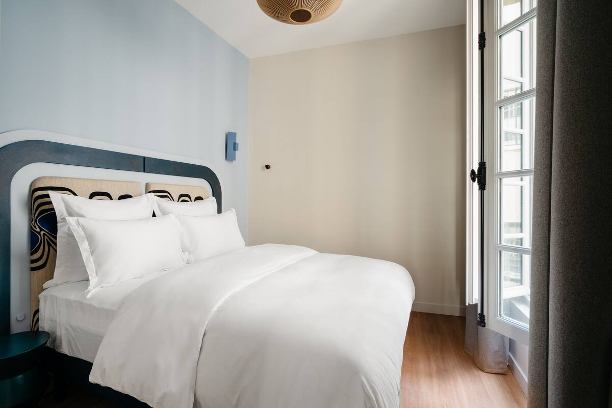 A neatly made bed with white linens in a room with light blue walls, a wooden headboard, and a window with curtains.