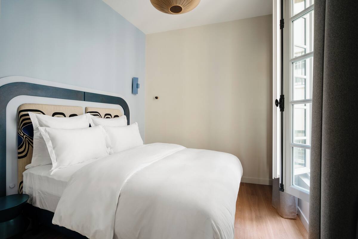 A neatly made bed with white linens in a room with light blue walls, a wooden headboard, and a window with curtains.