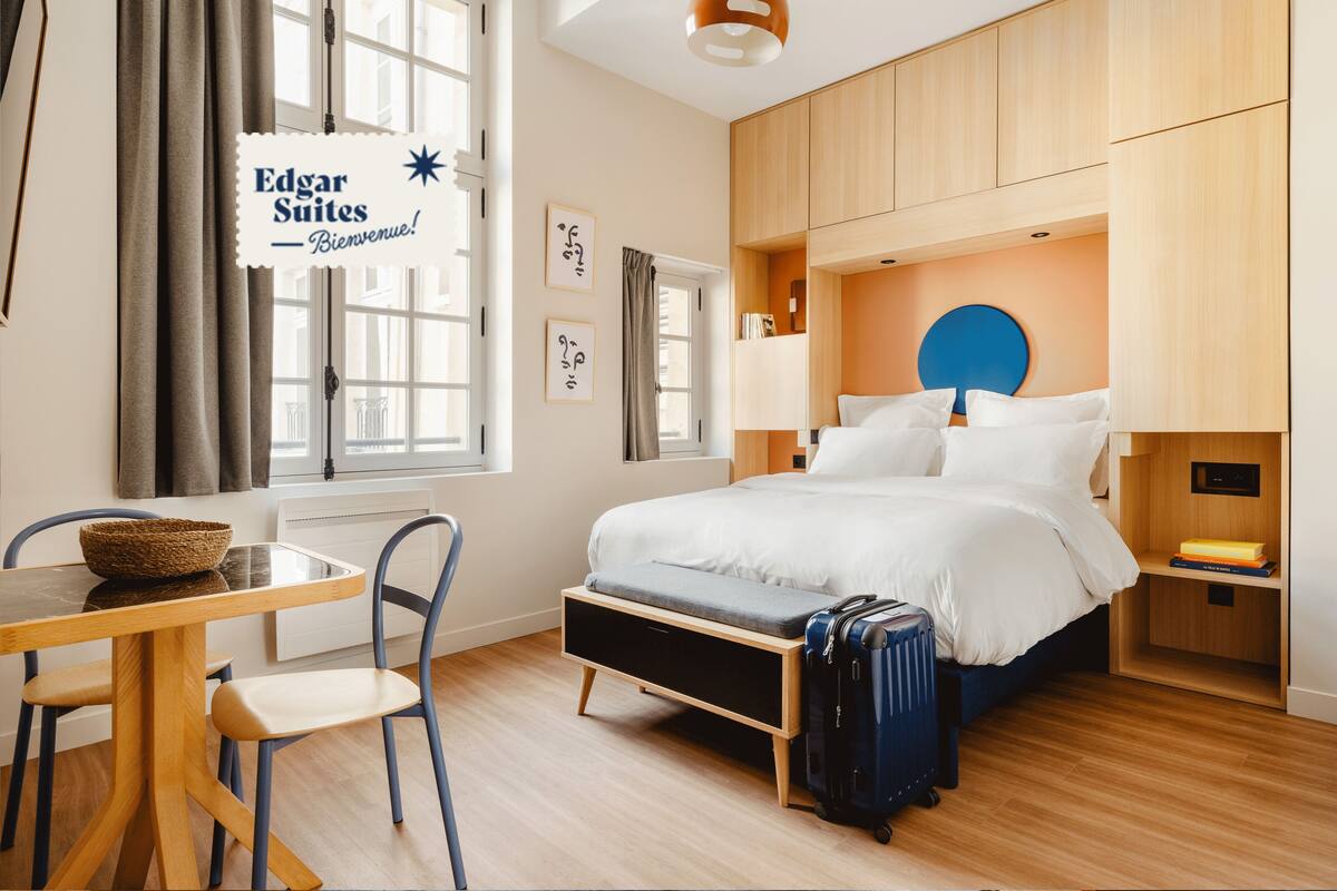 A hotel room with a bed, bedside table, chair, and a suitcase. There is a window with curtains, a wall-mounted sign, and a wall clock.
