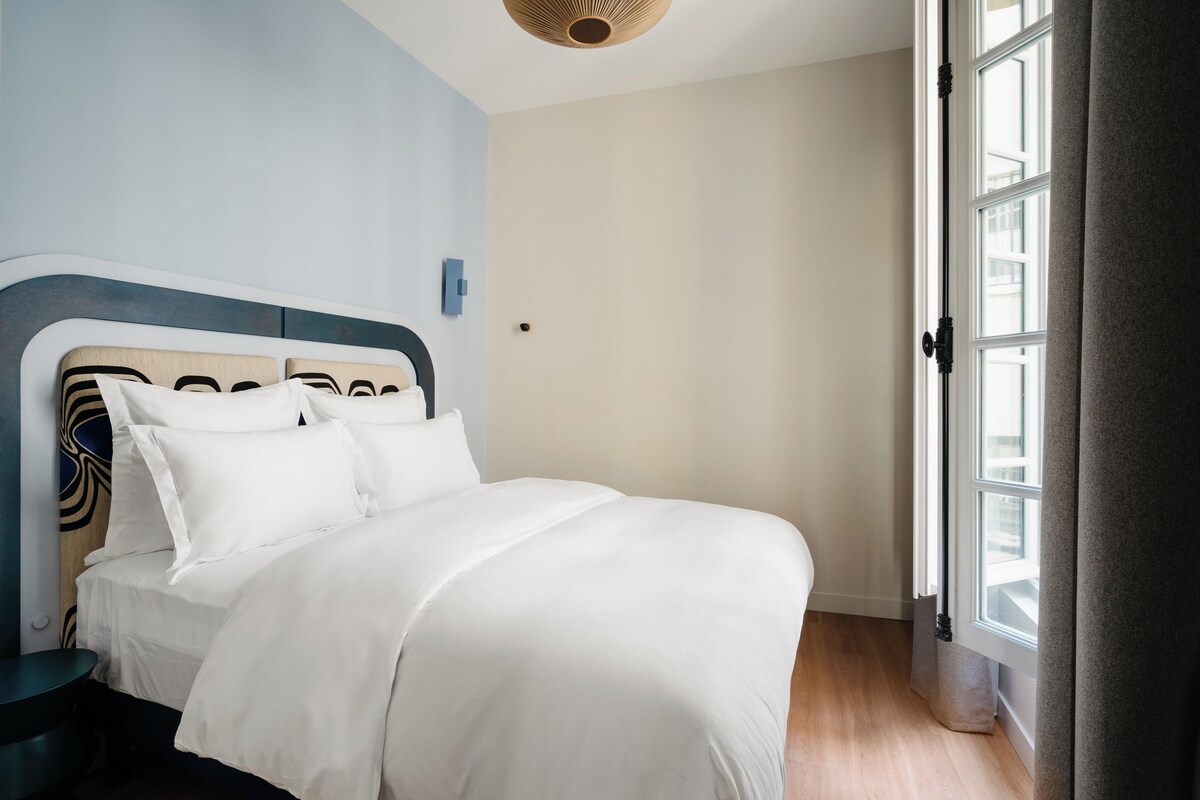 Una cama bien hecha en una habitación con un ventanal grande, un reloj de pared y una cortina en la puerta.