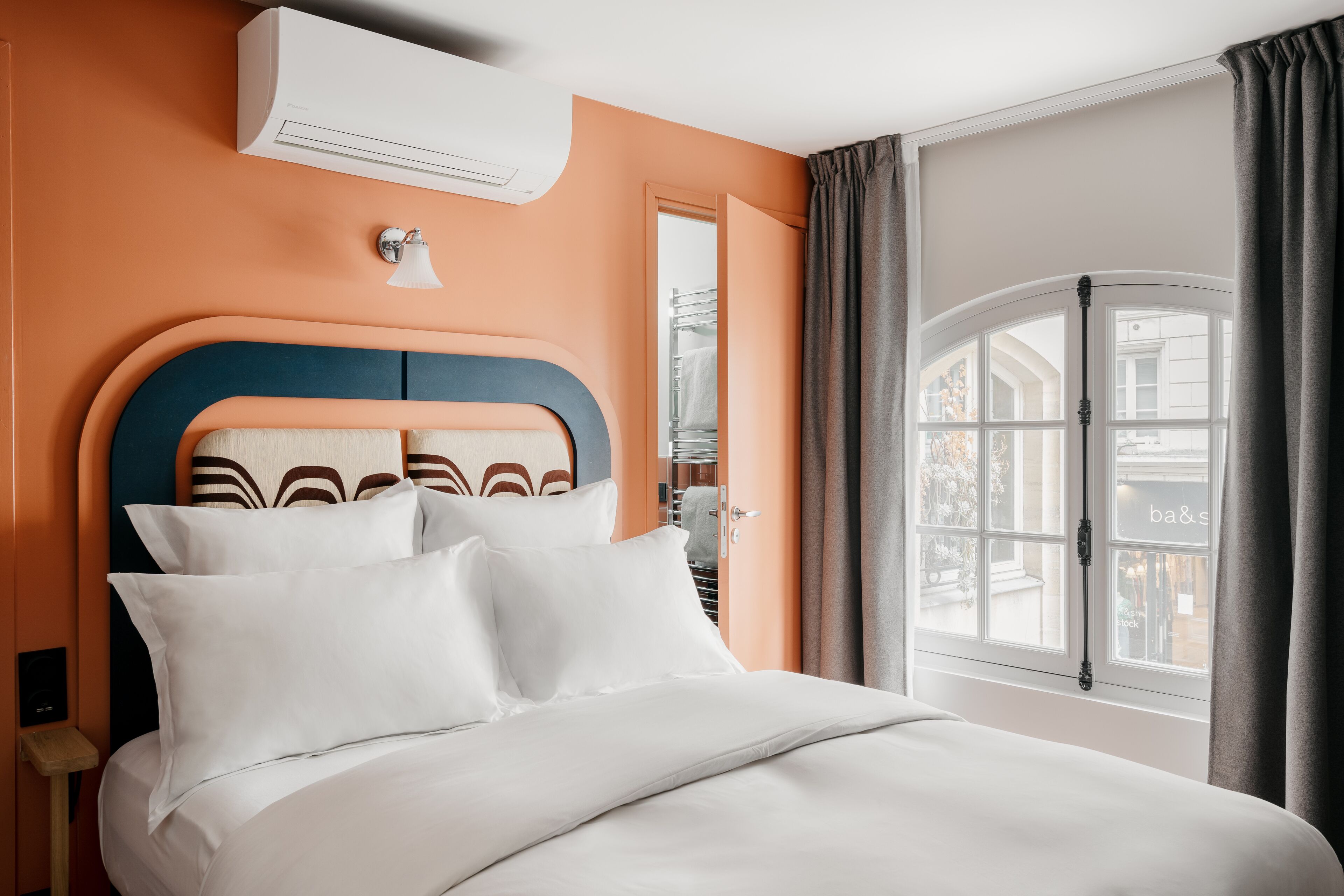 Habitación de hotel con cama, aire acondicionado, mesita de noche, espejo y ventana con cortinas.