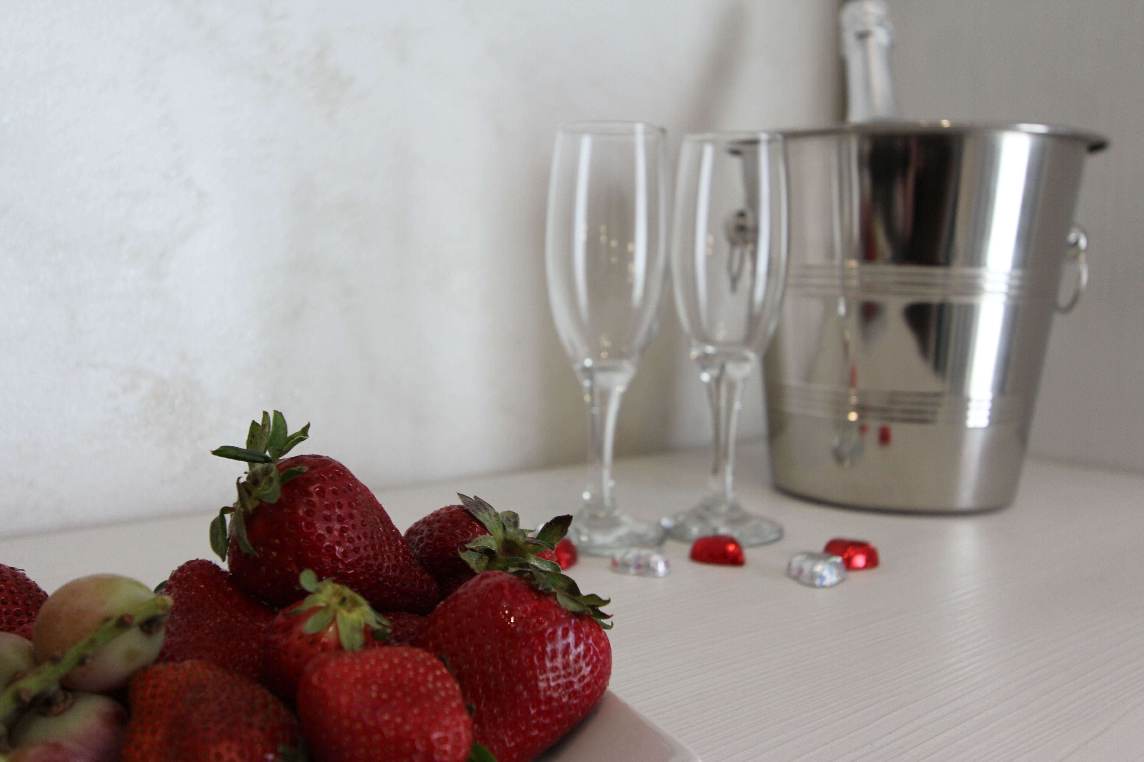 A white table with a plate of strawberries, two empty wine glasses, and a silver bucket.