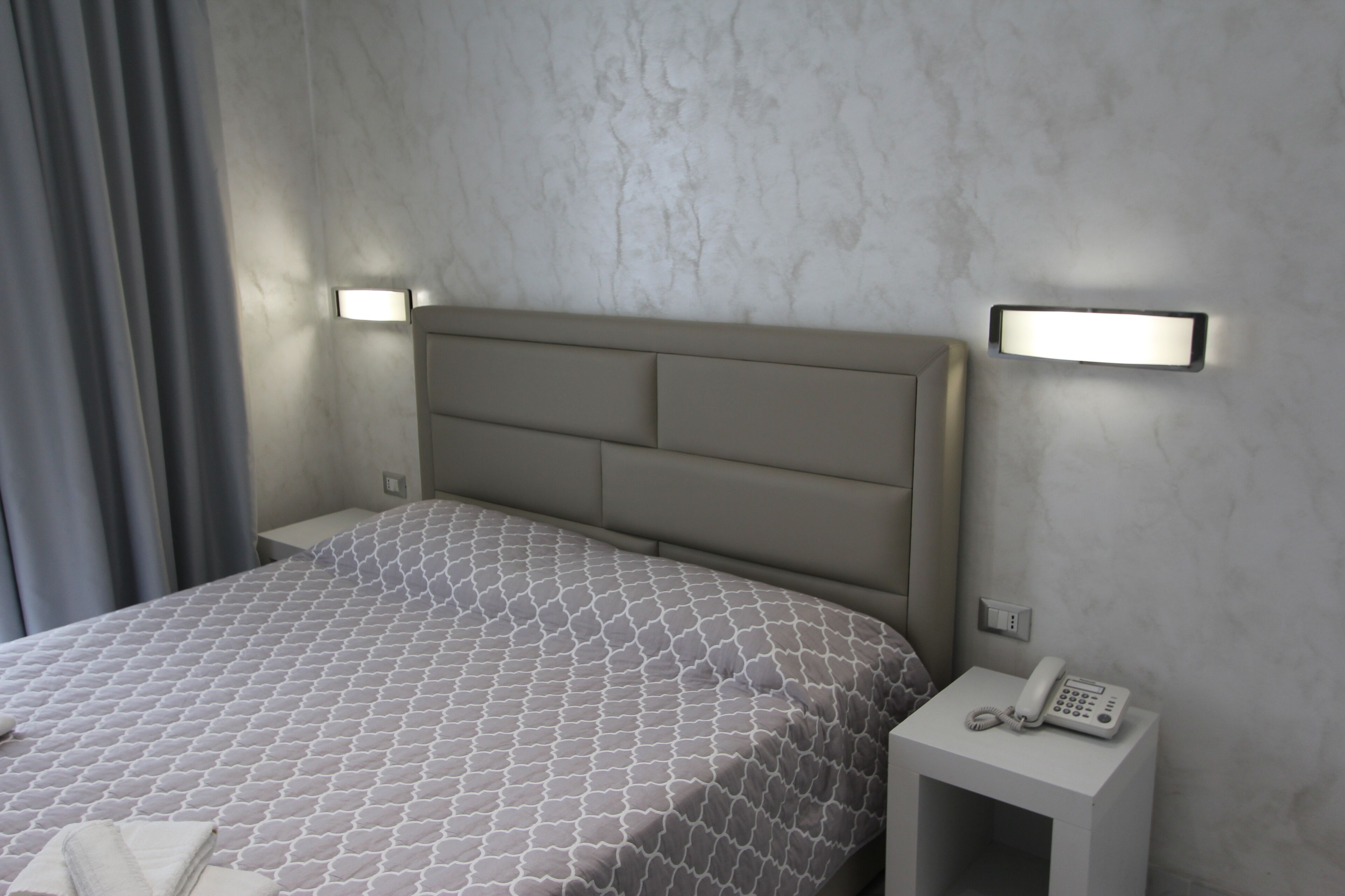 Una habitación de hotel con cama, mesita de noche con teléfono y luz de pared.