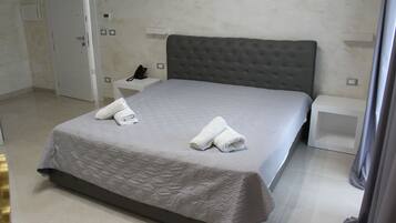 Un lit bien fait, avec une couverture grise et deux serviettes, une table de chevet avec un téléphone et une armoire blanche, dans une chambre d’hôtel.
