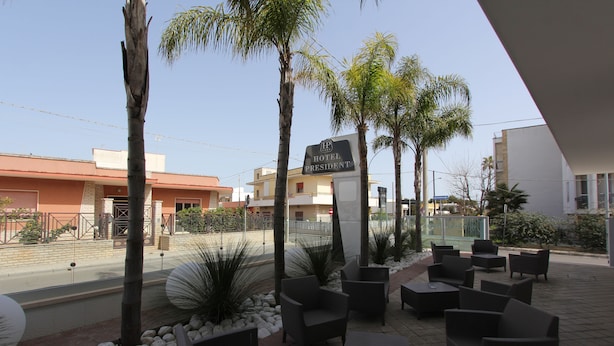 An outdoor seating area with palm trees, a sign reading "Hotel President", and a view of the street. - Hotel Presidente (Porto Cesareo)