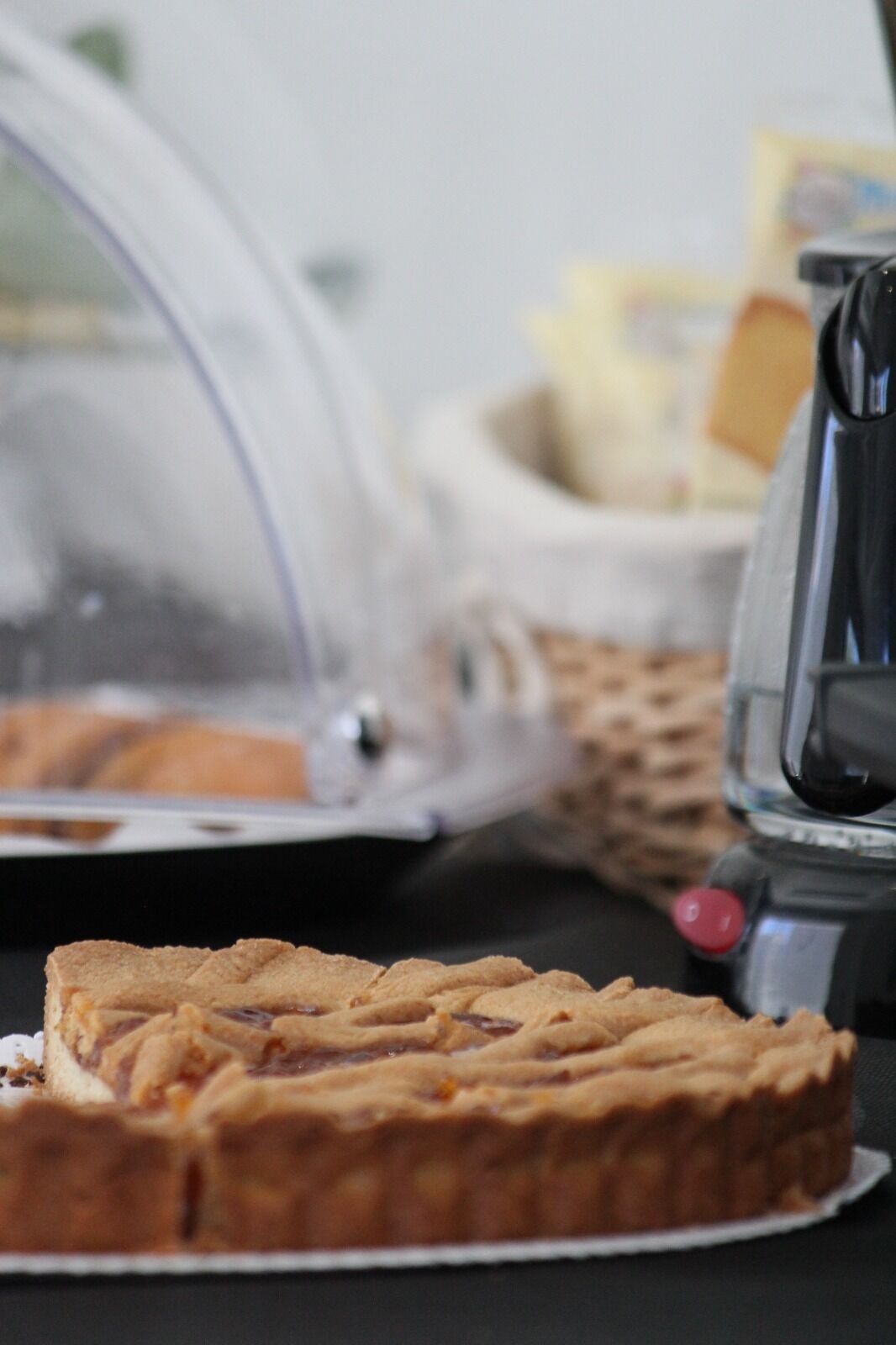 A pie with a lattice crust on a plate, a coffee machine, and a basket with bread in the background.
