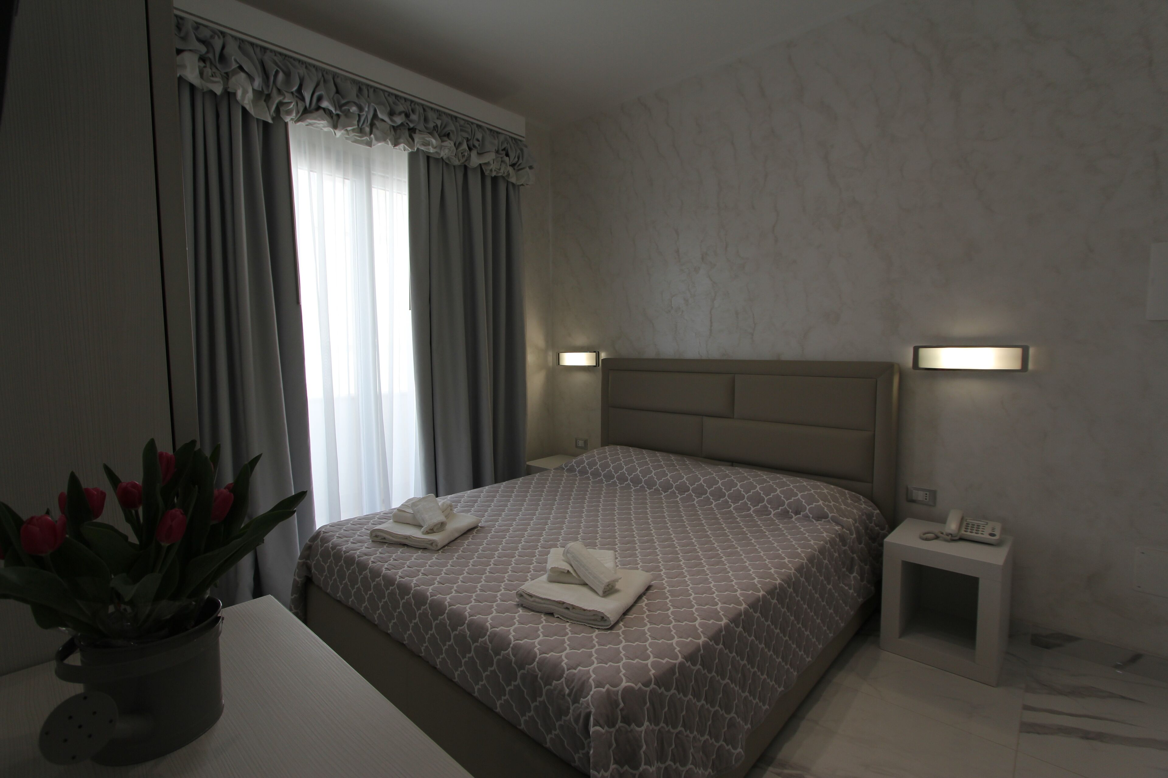 A hotel room with a bed, bedside tables, a potted plant, and a window with curtains.