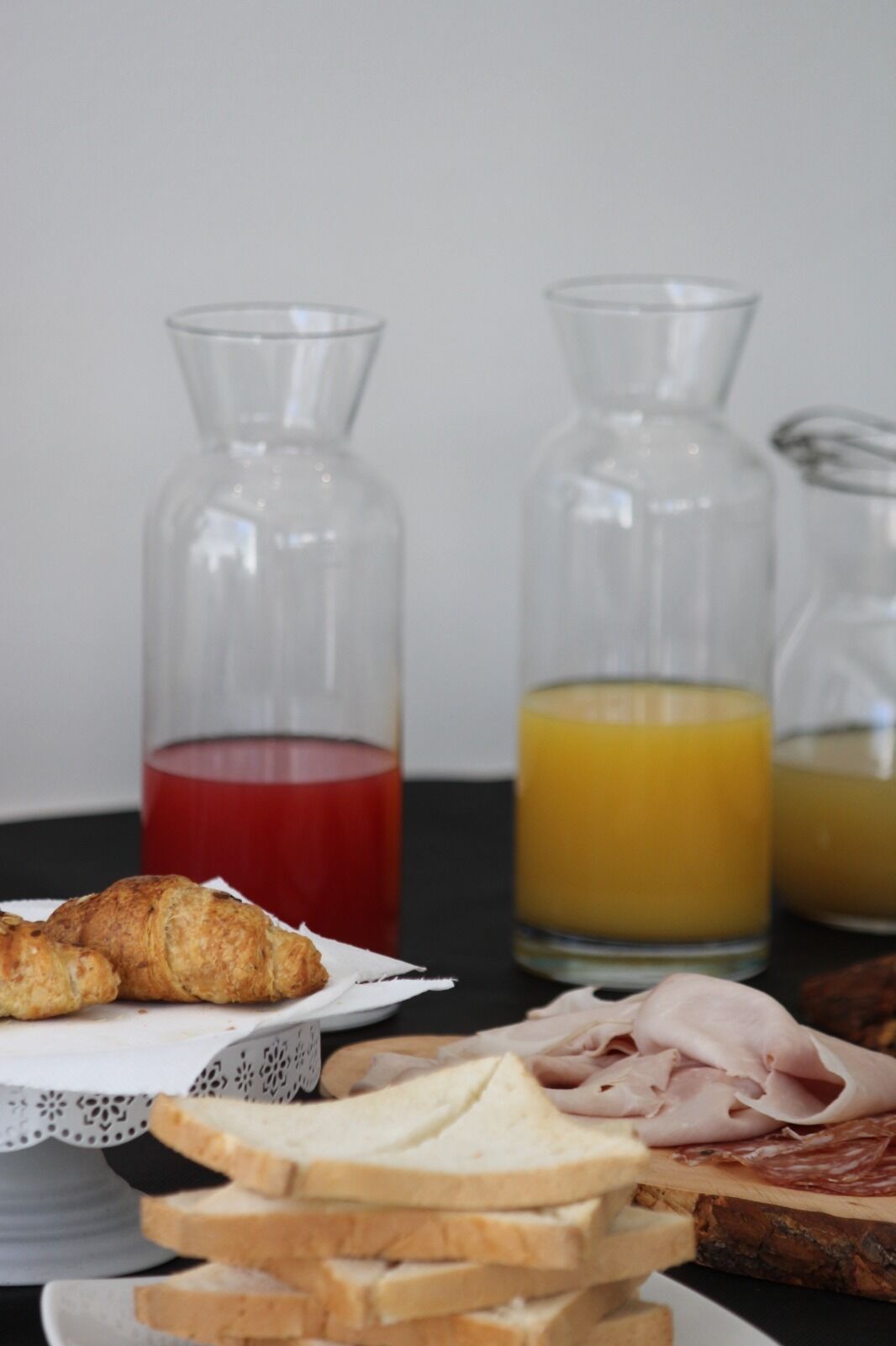 A breakfast table with croissants, sliced bread, and ham, along with three glass bottles containing red, yellow, and orange liquids.