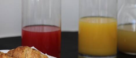 A breakfast table with croissants, sliced bread, and ham, along with three glass bottles containing red, yellow, and orange liquids.