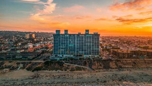 A modern building with a glass facade, situated on a coastal area with a view of the ocean and a sunset in the background. - Capri by the Sea 905 | Ocean View | Full Kitchen | Private Bedroom | Beach Access | On the Boardwalk (San Diego)