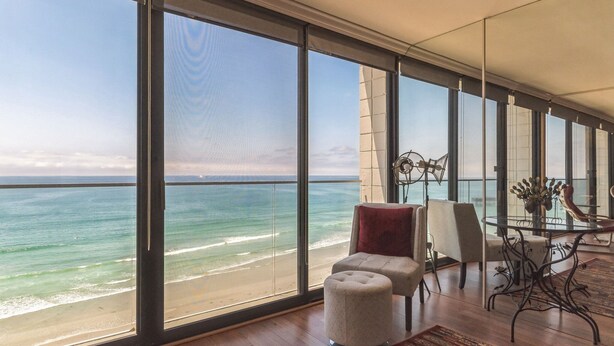 A spacious room with large windows overlooking a beach, a round table with chairs, and a small ottoman. - Capri by the Sea 905 | Ocean View | Full Kitchen | Private Bedroom | Beach Access | On the Boardwalk (San Diego)
