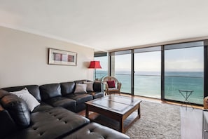 A modern living room with a black leather sofa, a wooden coffee table, and a view of the ocean through large windows. - Capri by the Sea 905 | Ocean View | Full Kitchen | Private Bedroom | Beach Access | On the Boardwalk (San Diego)