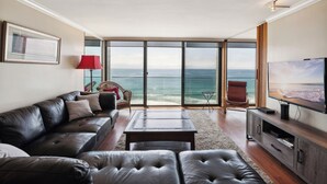 A living room with a leather sofa, a coffee table, a TV, and a view of the ocean through large windows. - Capri by the Sea 905 | Ocean View | Full Kitchen | Private Bedroom | Beach Access | On the Boardwalk (San Diego)