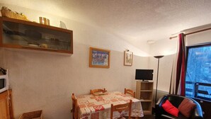 Un comedor acogedor con una mesa dispuesta para cuatro, un sofá de cuero negro con cojines rojos, un televisor sobre un mueble bajo y una ventana con vistas a árboles.