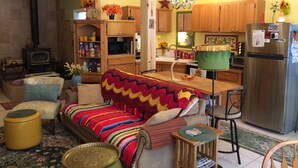 A cozy living room with a sofa, a coffee table, and a small wooden table with magazines. There is a kitchen area with wooden cabinets, a refrigerator, and a microwave.