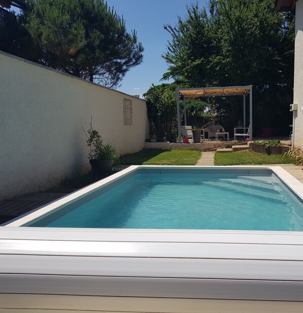 Une piscine rectangulaire bordée de blanc, entourée d’une zone pavée avec un petit jardin et une pergola en arrière-plan.