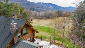 A log cabin with a stone chimney, a deck with outdoor furniture, and a view of a mountainous landscape. - Luxury Log Cabin-Windham Mountain.  Amazing sunsets, large lawn, deck, fireplace (Windham)