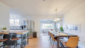 A modern kitchen with a central island, bar stools, and a dining area with chairs. - Huge Yard with Marsh Views! | Minutes to Beach/DT! (St. Augustine)