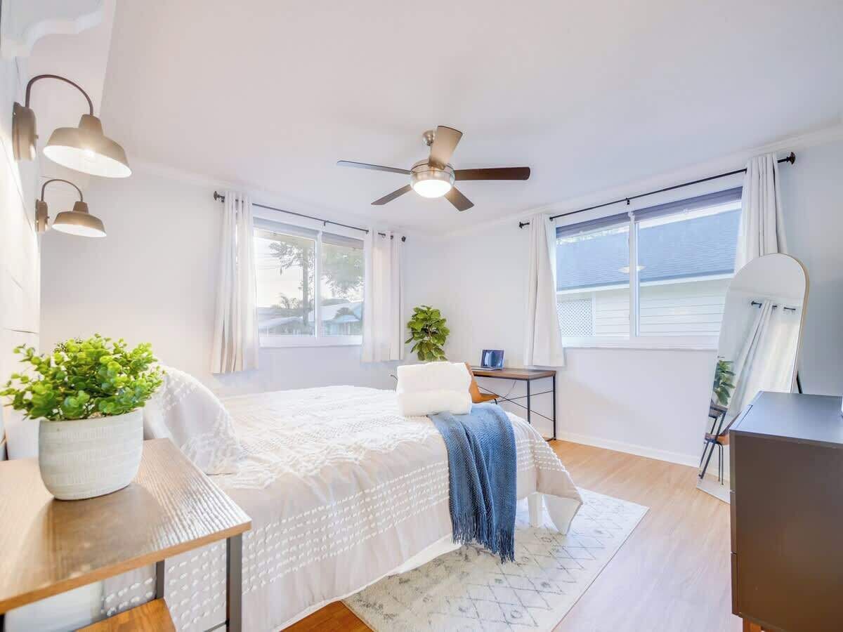 A bedroom with a bed, ceiling fan, large windows, and a desk with a plant.