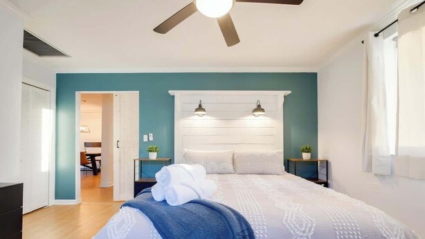 A bedroom with a bed, bedside tables, a ceiling fan, and a view into another room. - Huge Yard with Marsh Views! | Minutes to Beach/DT! (St. Augustine)