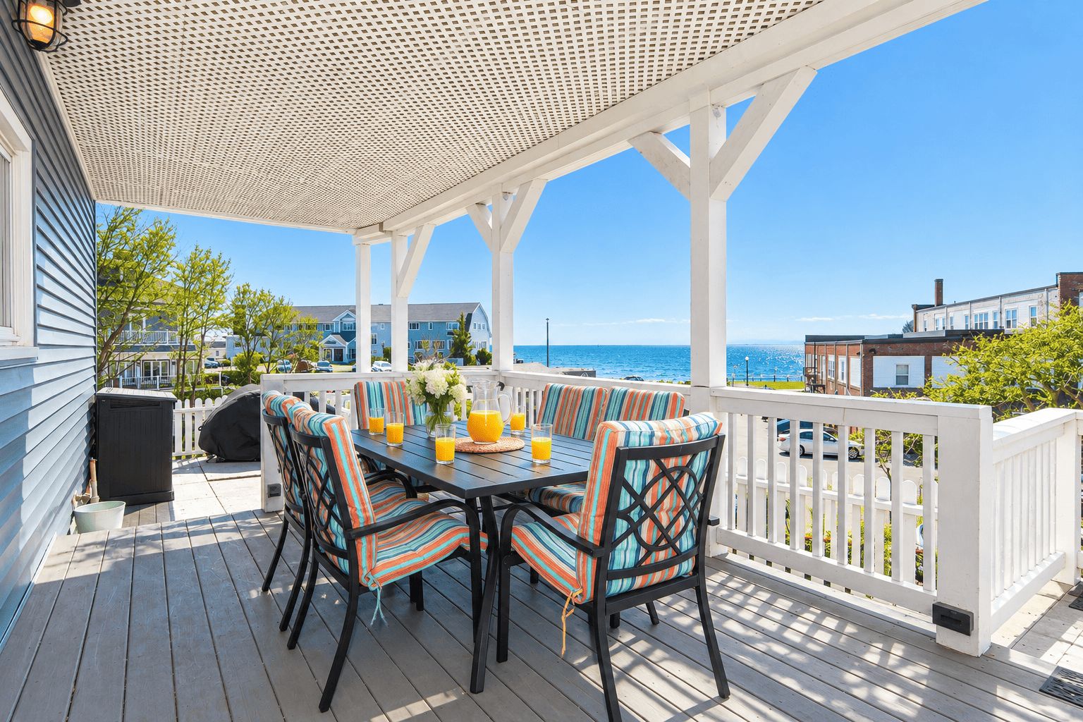Une terrasse extérieure couverte, avec une table dressée pour deux personnes, des chaises dotées de coussins à rayures et une vue sur l’océan.
