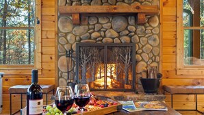 L’intérieur chaleureux d’un chalet avec une cheminée en pierre, des murs en bois et un coin repas agrémenté d’une bouteille de vin, de verres et d’un plateau de fruits.