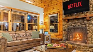 L’intérieur d’un chalet confortable, avec des murs en rondins de bois, une cheminée en pierre, un canapé en cuir, une table basse en bois sur laquelle est disposée une corbeille de fruits, et un téléviseur affichant le logo « NETFLIX ».