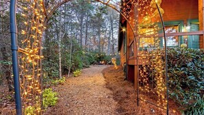 Une cabane en bois dotée d’un porche couvert, entourée d’une zone boisée et décorée de guirlandes lumineuses.