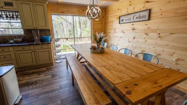 Interno di una baita in legno con cucina, tavolo da pranzo e sedute su panche.